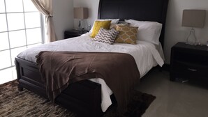 A bedroom with a large bed, a dark headboard, and multiple pillows. There are two bedside tables with lamps and a window with curtains. - Luxury Condo at Venito (Nassau)