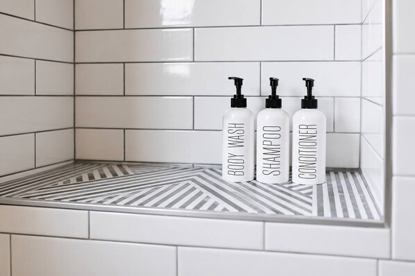 A bathroom with white tiled walls and a striped floor. There are three bottles labeled 'BODY WASH,' 'SHAMPOO,' and 'CONDITIONER' on a shelf.
