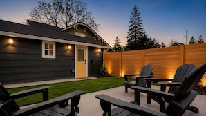 Una casa con fachada oscura, una puerta amarilla y una valla de madera. En un patio empedrado hay dos sillas Adirondack negras y una mesita.