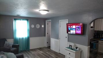 A living room with a flat-screen TV, a sofa, a coffee table, and a kitchen area with cabinets and appliances.