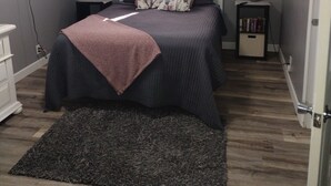 Ein Schlafzimmer mit einem Bett, einem an der Wand befestigten Fernseher, einem Fenster mit Vorhängen, einem Nachttisch mit einer Lampe und einem Bücherregal.