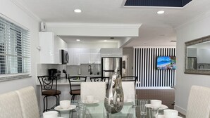 Un espace repas moderne avec une table en verre disposée pour six personnes, entourée de chaises blanches à revêtement. On aperçoit à l’arrière-plan une cuisine avec des armoires blanches et un réfrigérateur en acier inoxydable.