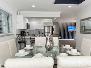 Un moderno comedor con una mesa de vidrio dispuesta para seis personas, rodeado de sillas blancas tapizadas. Se ve una cocina con armarios blancos y un refrigerador de acero inoxidable al fondo.