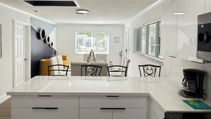 Une cuisine moderne dotée d’un îlot central, de meubles blancs et d’un coin repas avec une chaise jaune.