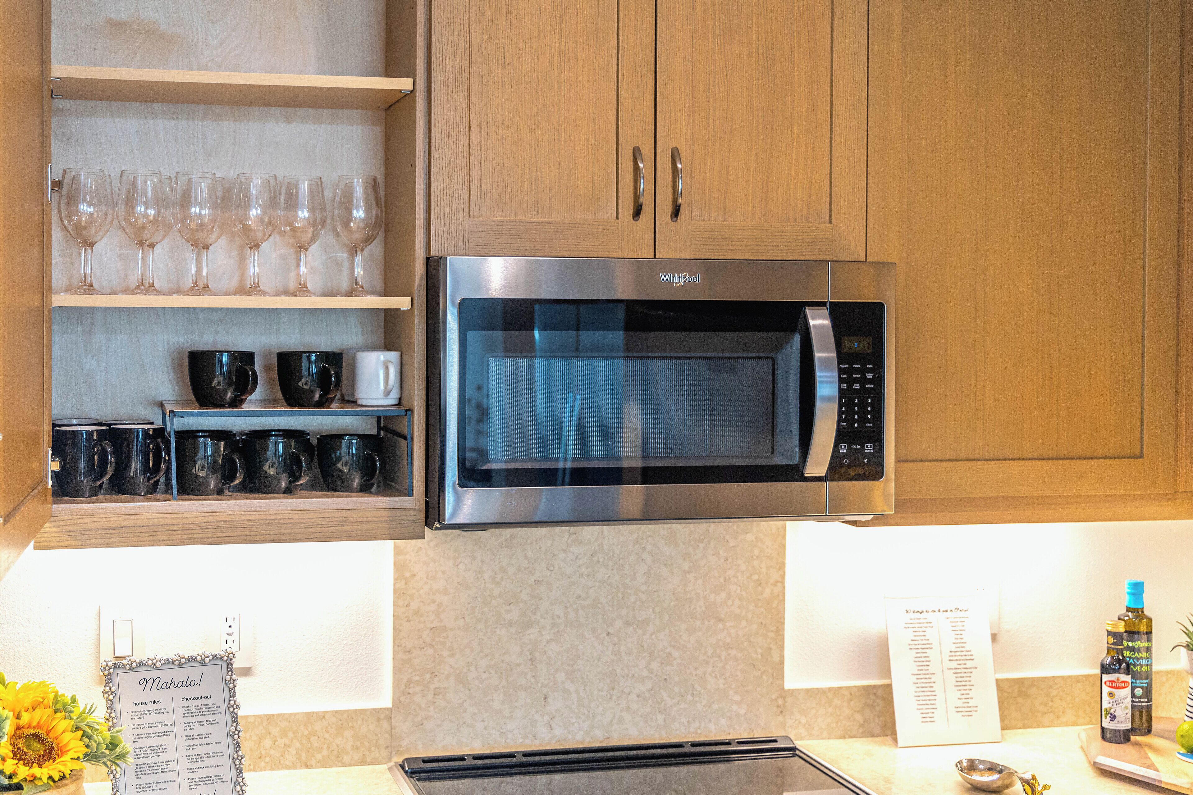 A kitchen with wooden cabinets, a stainless steel microwave, and shelves with wine glasses and black mugs.