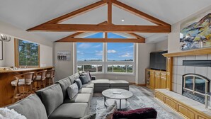 A spacious living room with a large sectional sofa, a round coffee table, and a fireplace. There is a bar area with wooden chairs and a large window offering a view of the outdoors. - Renovated home w/ breathtaking views of the bay and new hot tub (Bodega Bay)
