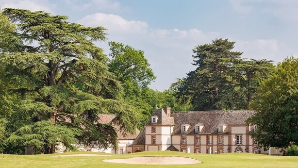 A large, historic building with multiple windows and a symmetrical facade, surrounded by lush greenery and a well-maintained lawn.