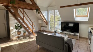 A spacious loft with a high ceiling, a wooden staircase, a grey sofa, a television, and large windows offering views of the outdoors.