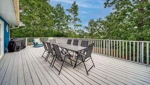 A covered outdoor deck with a table and chairs, surrounded by a white railing and trees.