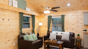Ein gemütliches Blockhaus-Interieur mit zwei Ledersofas, einem Holztisch, einem Deckenventilator und einem kleinen Schrank.