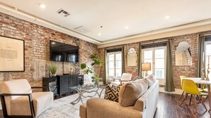 A living room with a brick wall, a sofa, a coffee table, and a dining area with a yellow chair. - 3B/2B The Jackson - 60 Day Min. Rental (New Orleans)