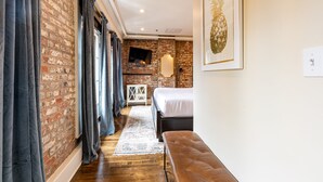 A bedroom with a brick wall, a bed, a bench, a TV, and a framed picture. - 3B/2B The Jackson - 60 Day Min. Rental (New Orleans)