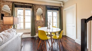 A dining area with a round table and yellow chairs, a brick wall, and a large window with curtains. - 3B/2B The Jackson - 60 Day Min. Rental (New Orleans)