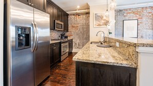 A modern kitchen with dark wood cabinets, stainless steel appliances, a granite countertop, and a brick accent wall. - 3B/2B The Jackson - 60 Day Min. Rental (New Orleans)