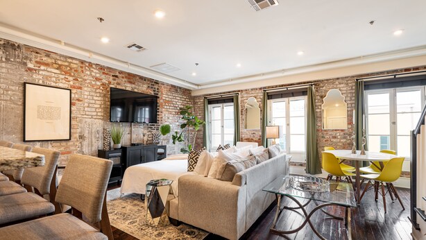 A modern room with a brick wall, a bed, a sofa, a dining table with chairs, and a TV mounted on the wall. - 3B/2B The Jackson - 60 Day Min. Rental (New Orleans)