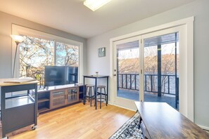 Una sala de estar moderna con piso de madera, un televisor, una mesita baja con taburetes y un amplio ventanal corredero que da a una terraza.
