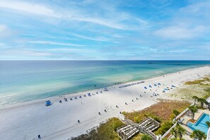 A beach with a pier extending into the ocean, a sandy beach with umbrellas, and a pool area.