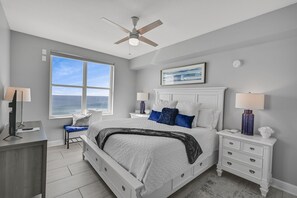 A bedroom with a bed, nightstands, a desk, a chair, a ceiling fan, and a large window with a view of the ocean.
