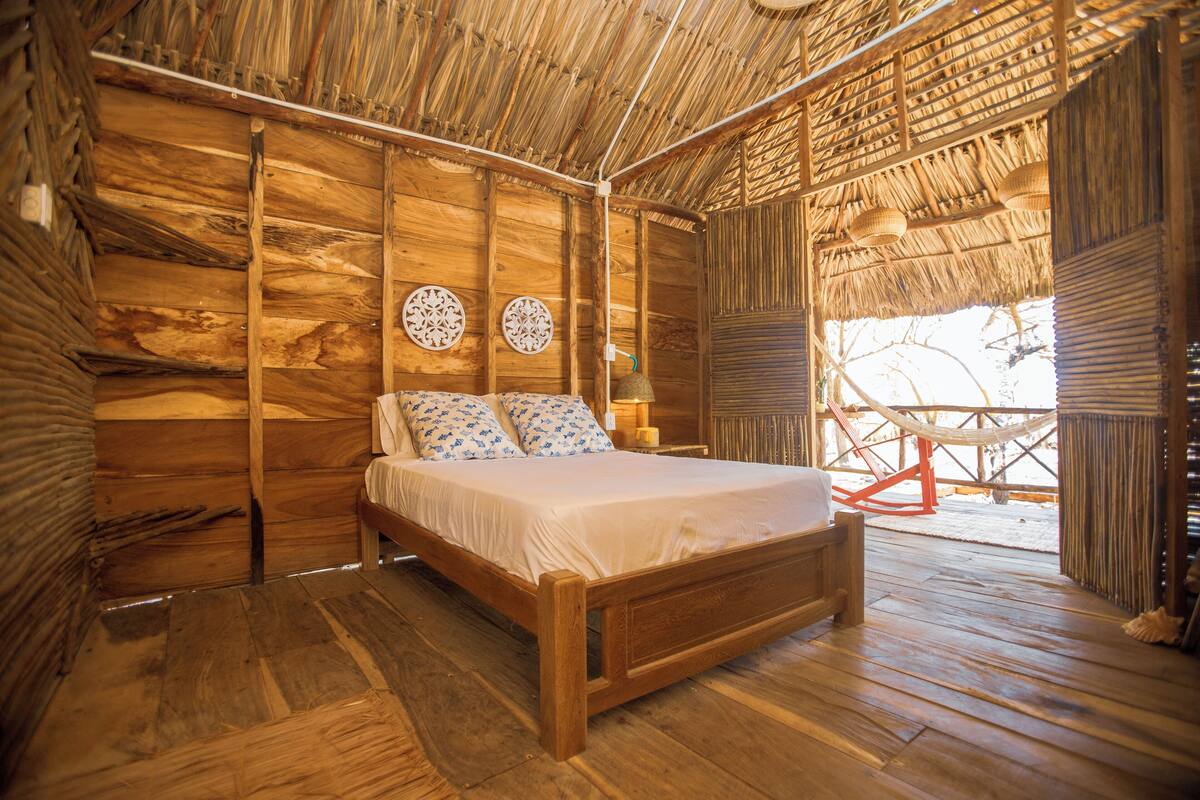 Ein Zimmer mit Holzboden, einem Bett, zwei Wandbildern und einem Strohdach.
