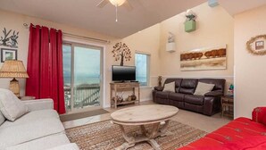 A living room with a sofa, a coffee table, a TV, and a balcony with red curtains. - Ocean Reef 701 3 Bedroom Townhouse by RedAwning (Gulf Shores)