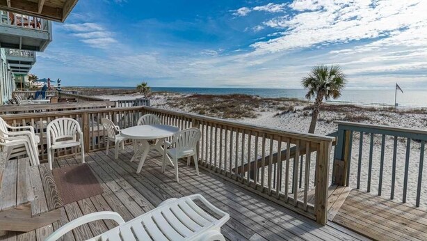 A wooden deck with white chairs and a table, overlooking a beach with sand dunes and a palm tree. - Ocean Reef 701 3 Bedroom Townhouse by RedAwning (Gulf Shores)