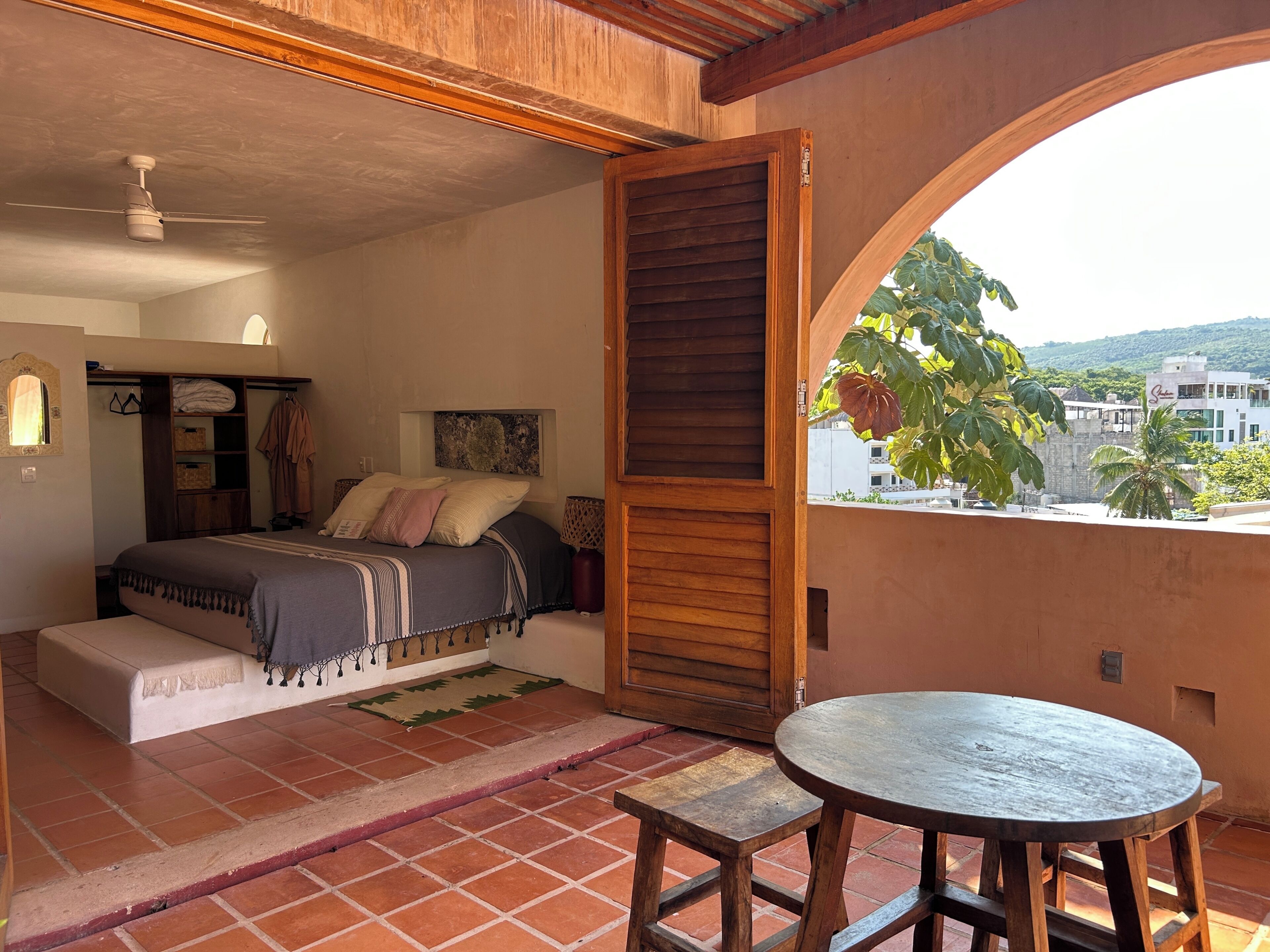 A bedroom with a bed, a wooden wardrobe, a small table, and a view through an open door.