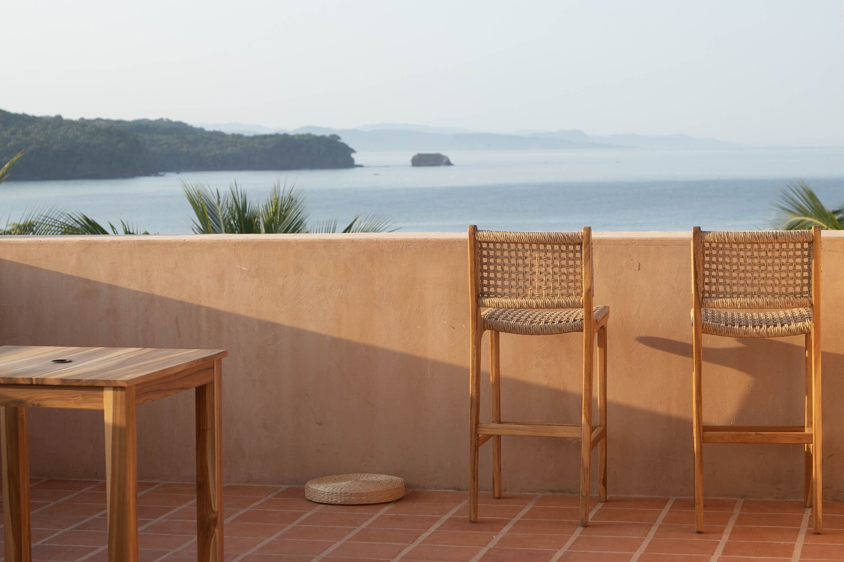 A terrace with two wicker chairs and a wooden table, overlooking a calm sea and distant hills.