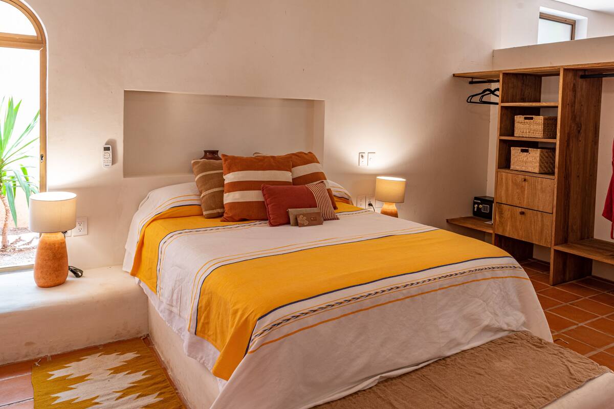 Une chambre à coucher avec un lit, des lampes de chevet, une armoire en bois et une fenêtre agrémentée d’une plante.