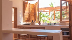 Una cocina con encimera blanca, taburetes de madera y vistas al exterior a través de amplios ventanales.