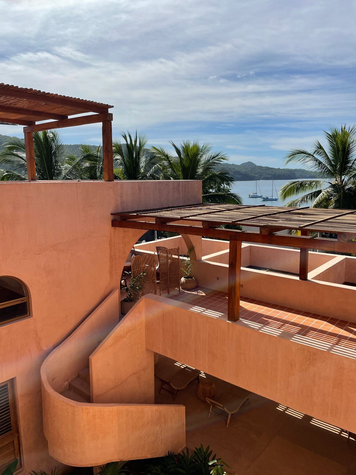 A modern outdoor patio with a wooden pergola, a curved staircase, and a view of the ocean with boats.