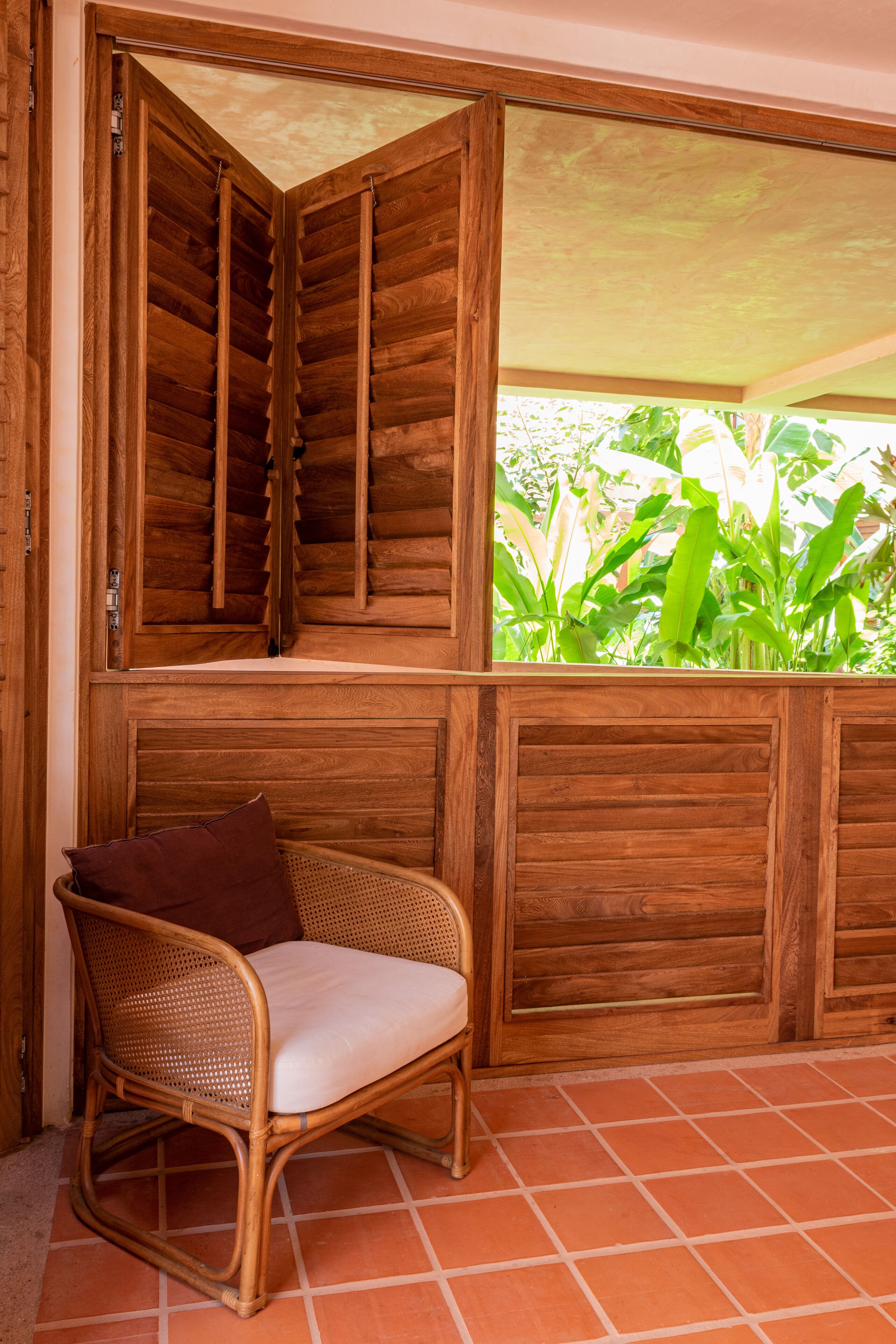 A wicker chair with a cushion in a room with wooden paneling and a window overlooking greenery.