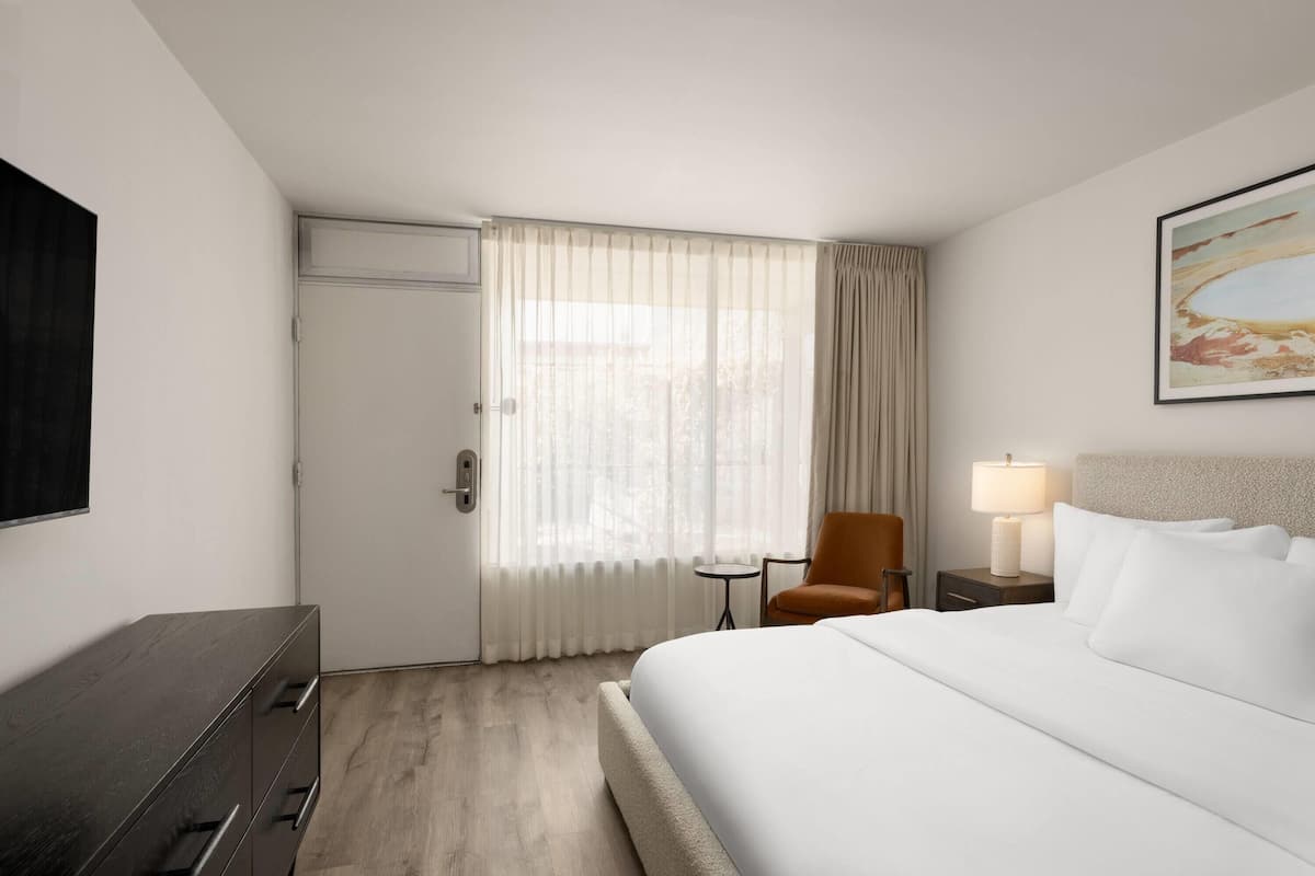 A hotel room with a bed, a chair, a desk, a lamp, and a painting on the wall.