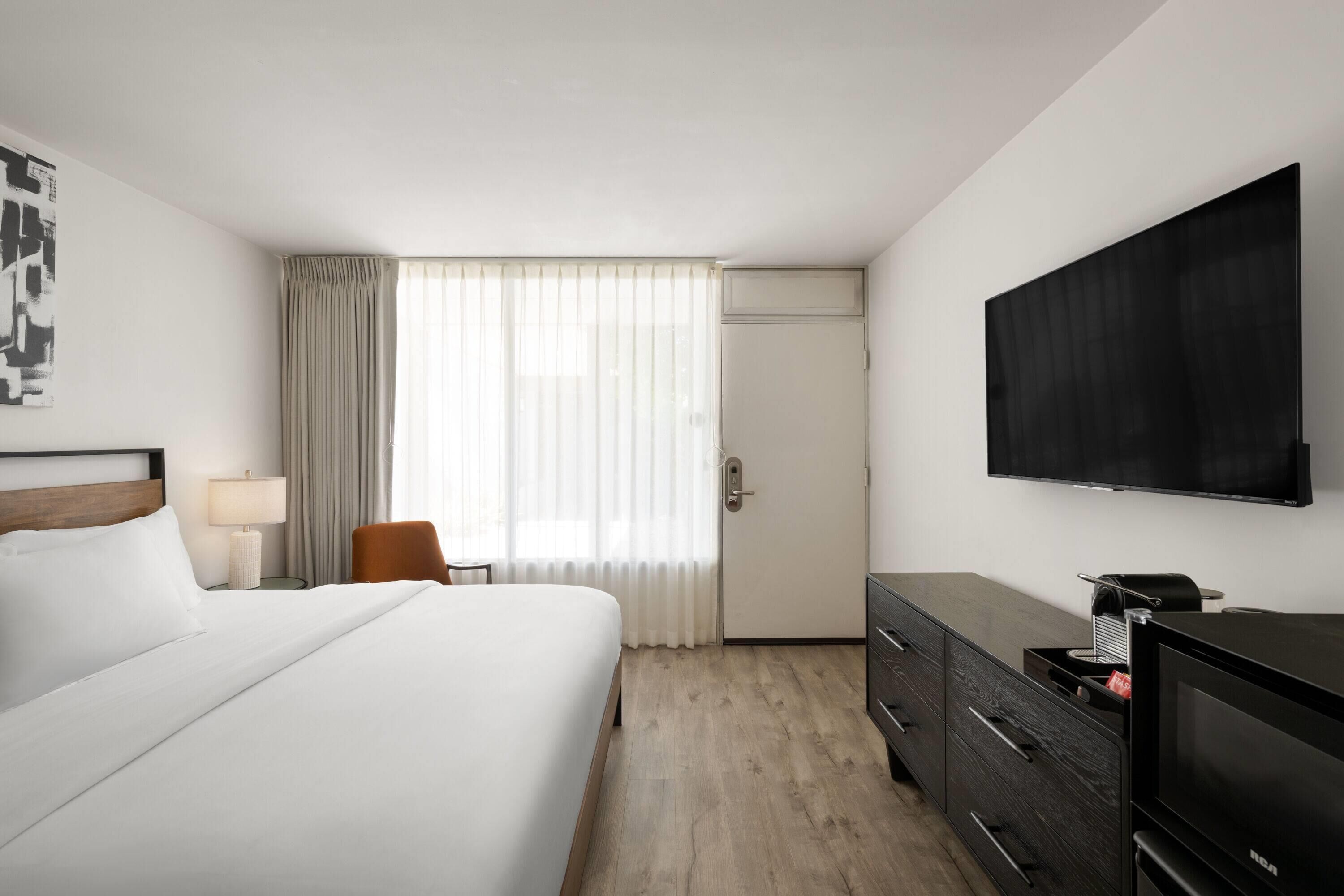 A hotel room with a large bed, a flat-screen TV, a desk with a lamp, and a window with curtains.