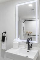 Un baño con lavamanos blanco, un espejo con luz, un perchero para toallas y una cortina de ducha.