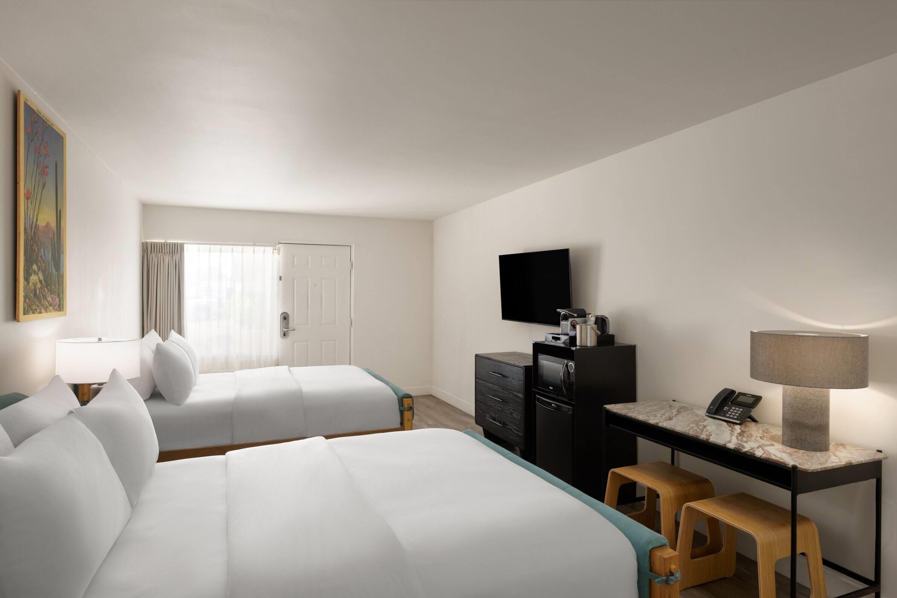 A hotel room with two beds, a desk with a lamp, a television, and a small stool.