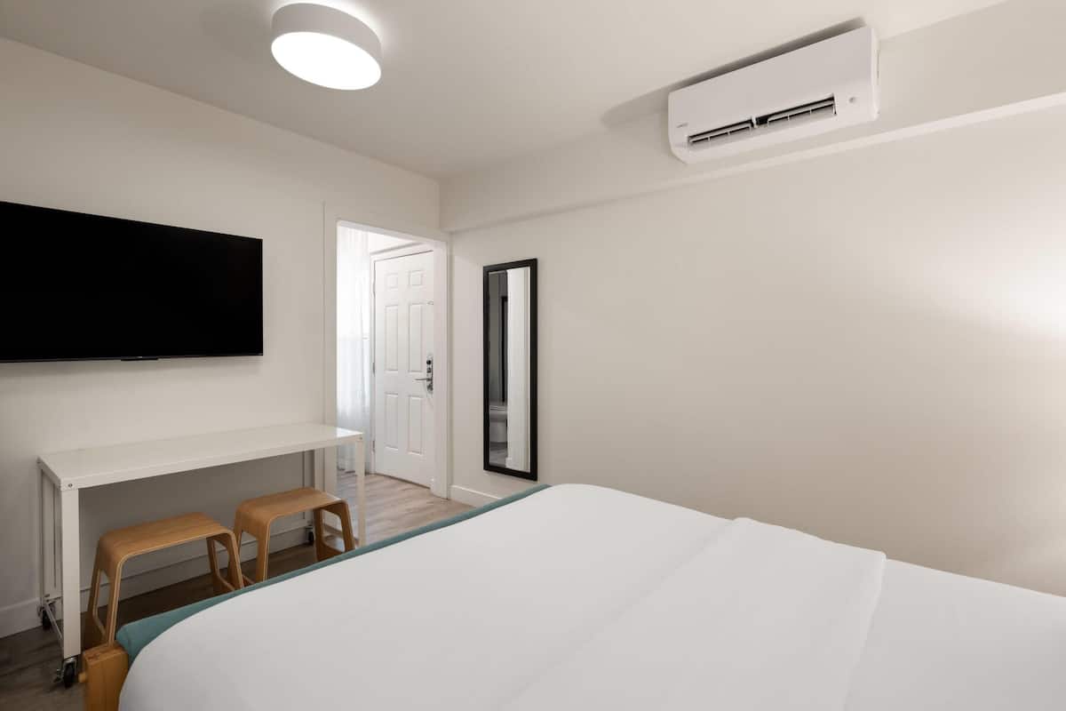 A hotel room with a bed, a desk, a chair, a TV mounted on the wall, and an air conditioning unit.