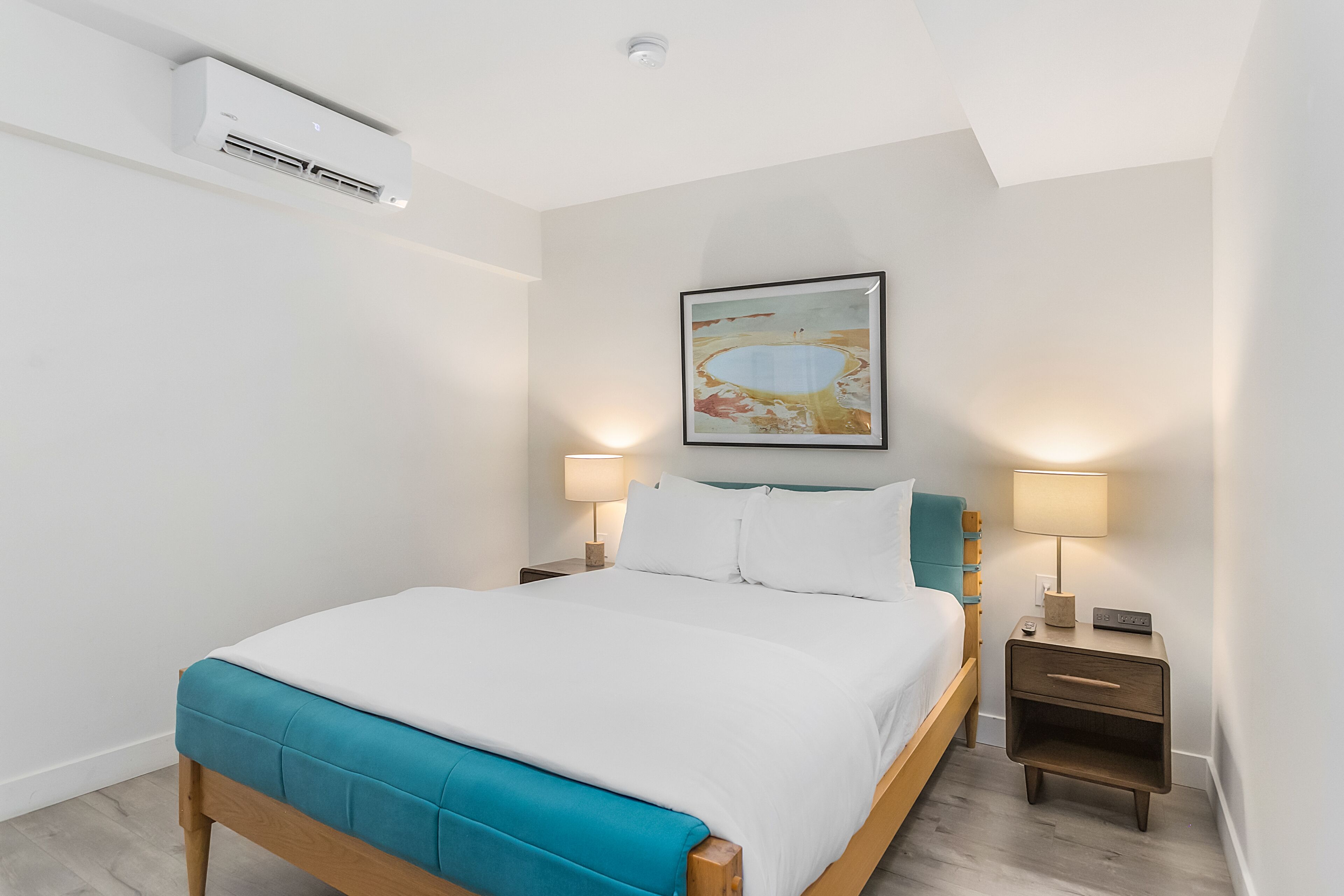 A neatly made bed with a teal accent, a bedside lamp, a nightstand, and a wall-mounted air conditioner.