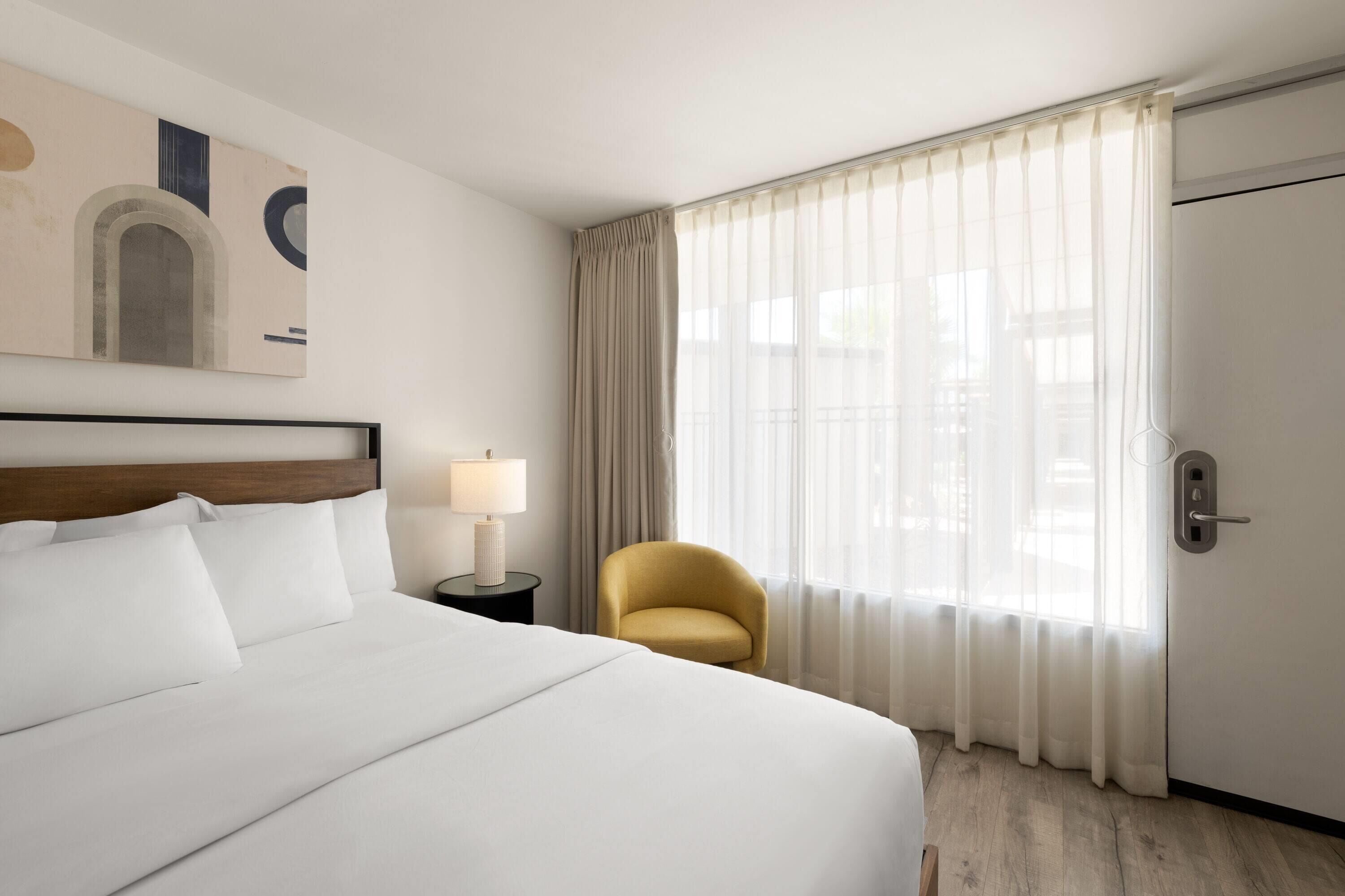 Une chambre d’hôtel avec un grand lit, une chaise, une table de chevet avec une lampe, une porte et une fenêtre avec des rideaux.