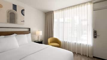A hotel room with a large bed, a chair, a nightstand with a lamp, a door, and a window with curtains.