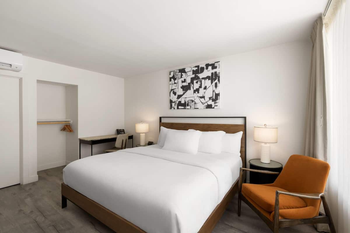 A hotel room with a large bed, two bedside tables with lamps, an orange chair, a wall art piece, and a door leading to another room.