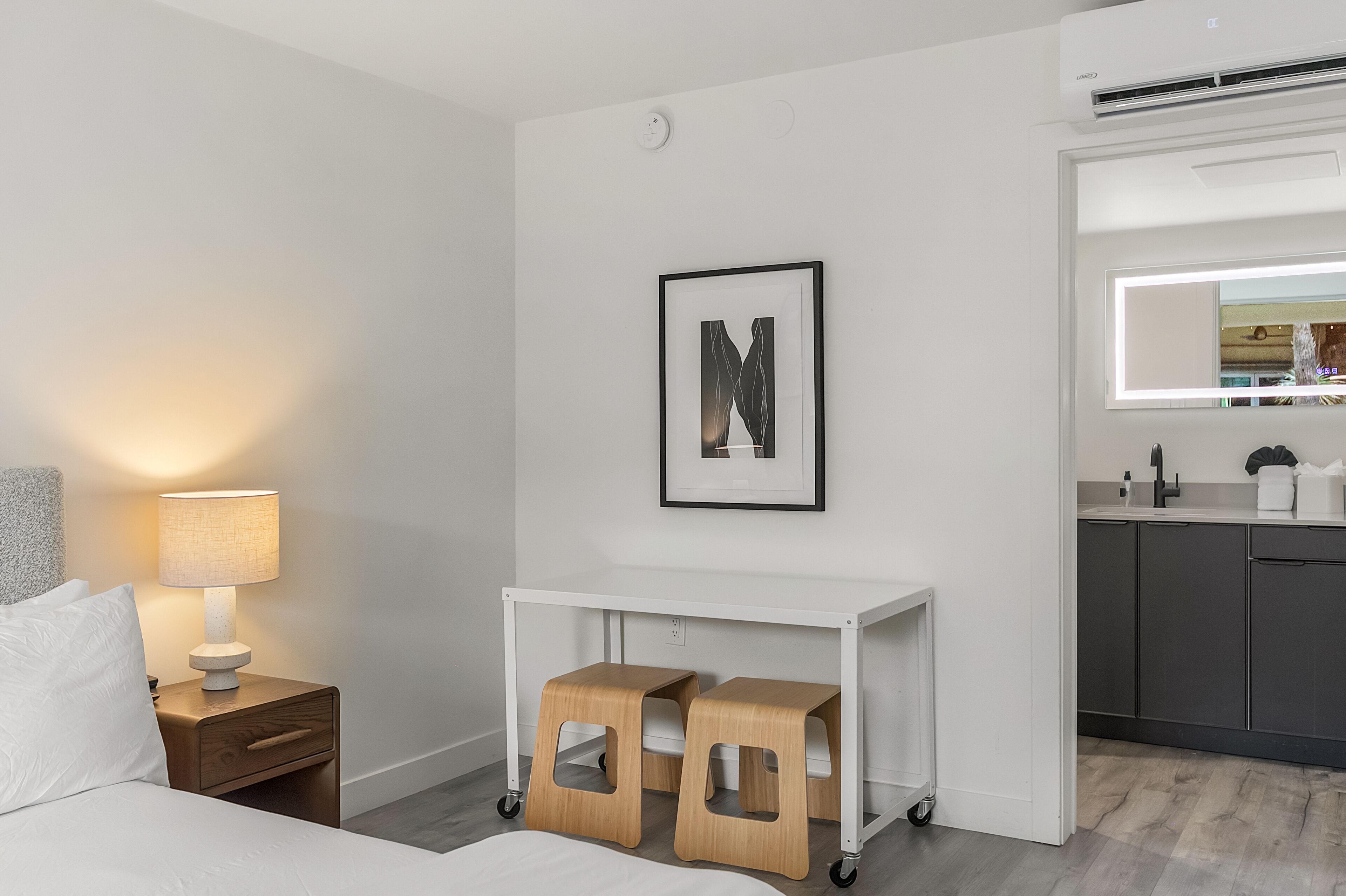 Une chambre d’hôtel comprenant un lit, une table de chevet avec une lampe, un bureau avec des tabourets, une lettre encadrée accrochée au mur et un coin cuisine avec des placards et un miroir.