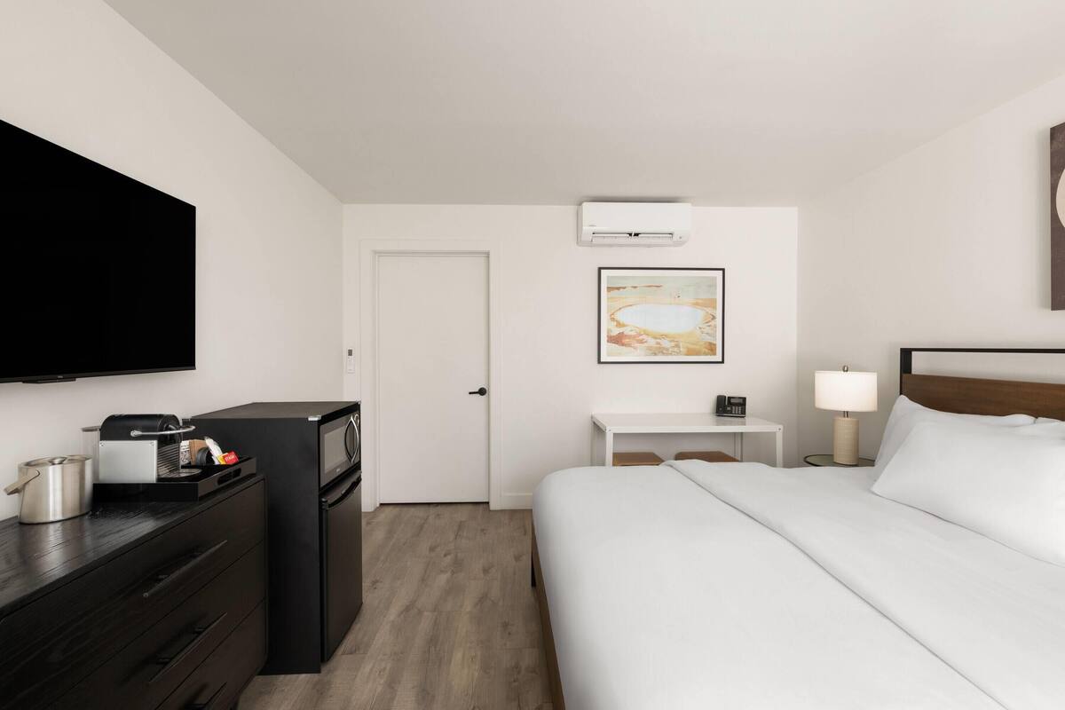 A hotel room with a bed, a desk, a flat-screen TV, a refrigerator, a microwave, a coffee maker, a lamp, and a framed picture on the wall.
