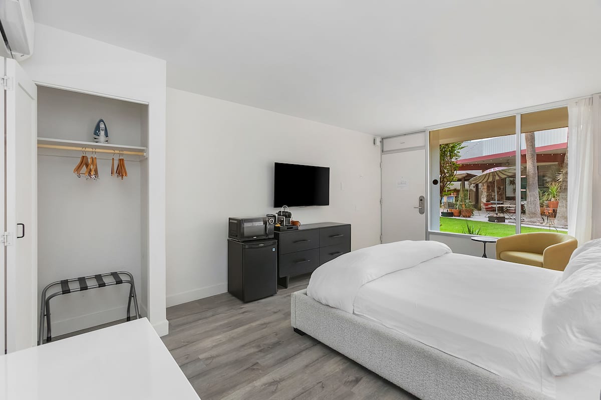 A hotel room with a bed, a desk, a wardrobe, a TV, and a view of an outdoor area with tables and chairs.