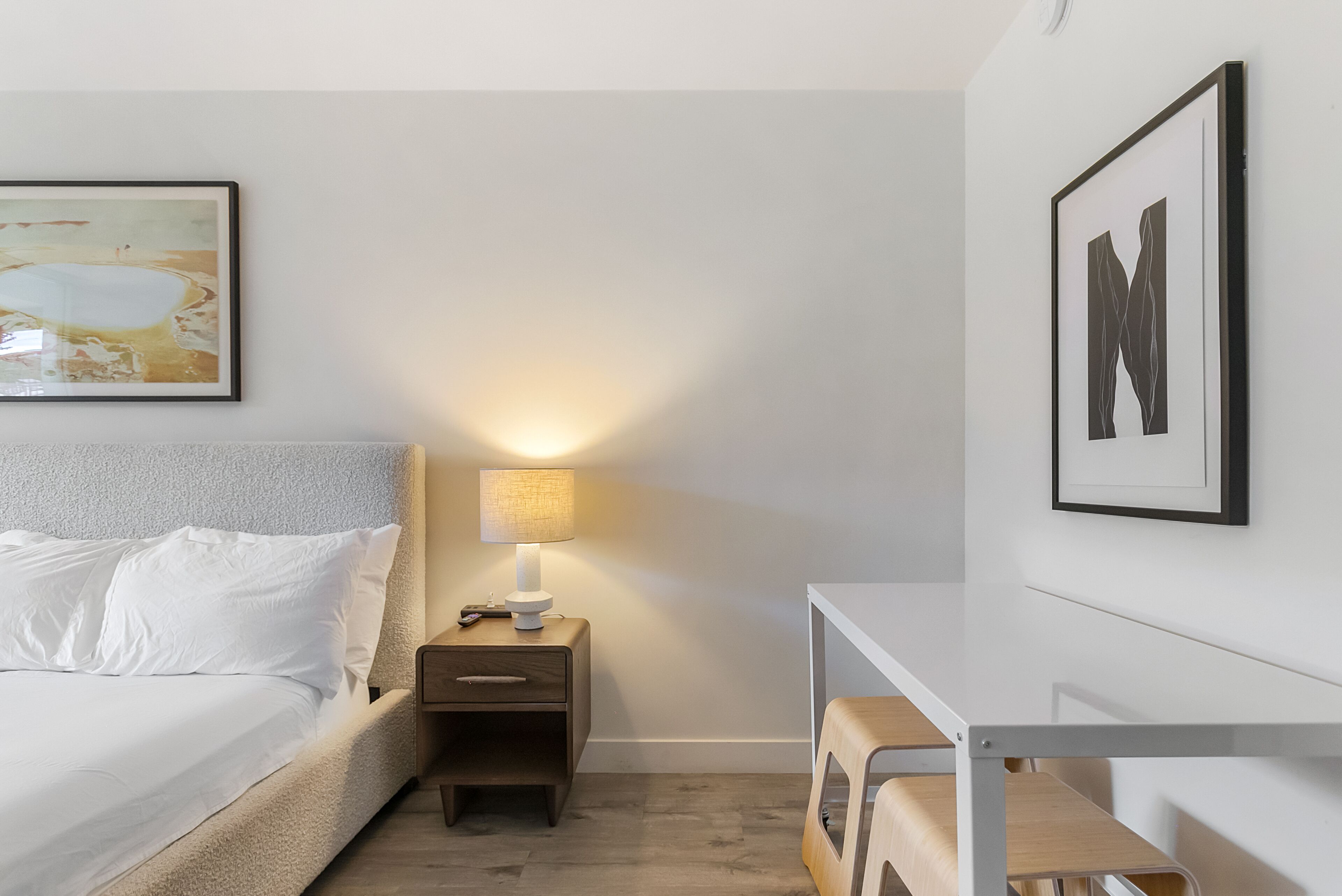 Un dormitorio con una cama, una mesita de noche con lámpara, un taburete de madera pequeño y dos cuadros enmarcados en la pared.