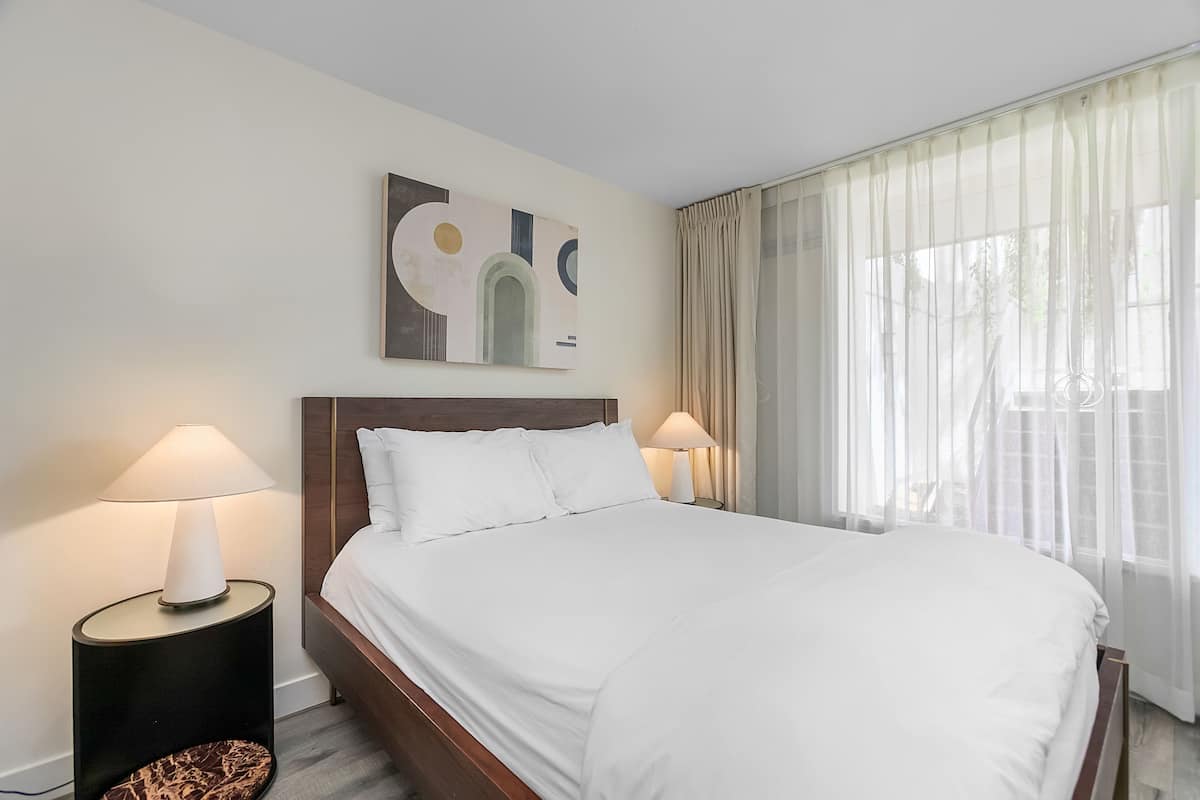A hotel room with a large bed, two bedside lamps, a headboard, and a window with sheer curtains.