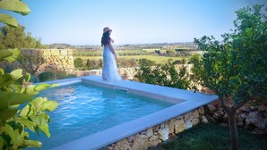 A woman in a white dress standing by a swimming pool with a stone wall and a view of a green landscape.
