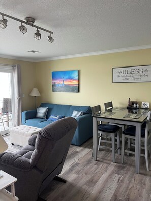 A living room with a blue sofa, a grey armchair, a dining table with chairs, a painting on the wall, and a ceiling light fixture.