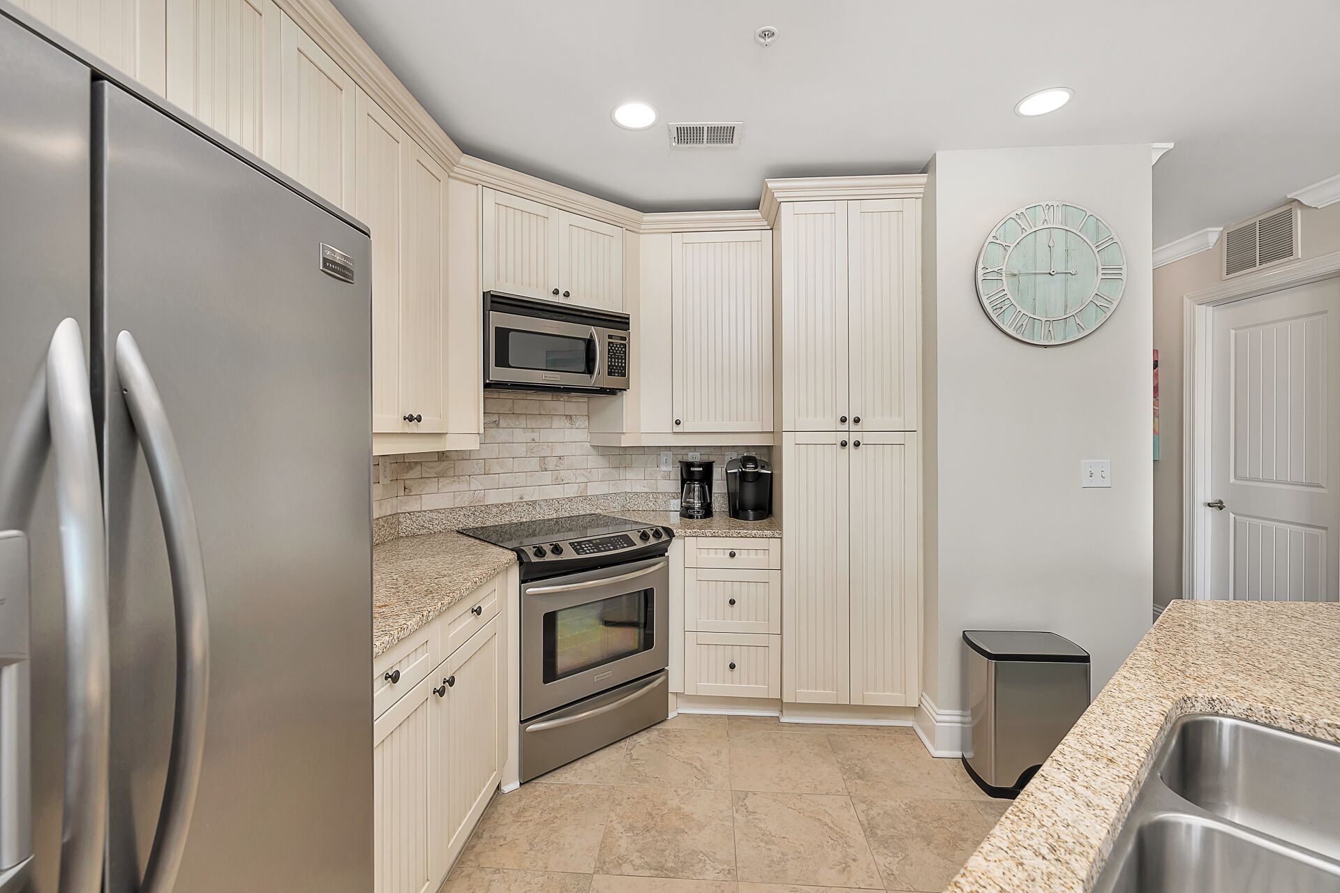 A modern kitchen with white cabinetry, stainless steel appliances, a granite countertop, and a clock on the wall.