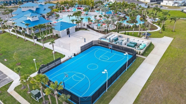 An aerial view of a resort with a large blue tennis court, multiple swimming pools, and surrounding palm trees.