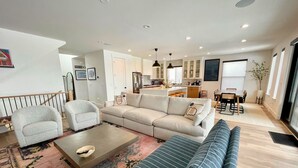 Una sala de estar moderna con un sofá, sillones y una mesita de centro. Se observa una cocina al fondo con armarios y una zona de comedor.